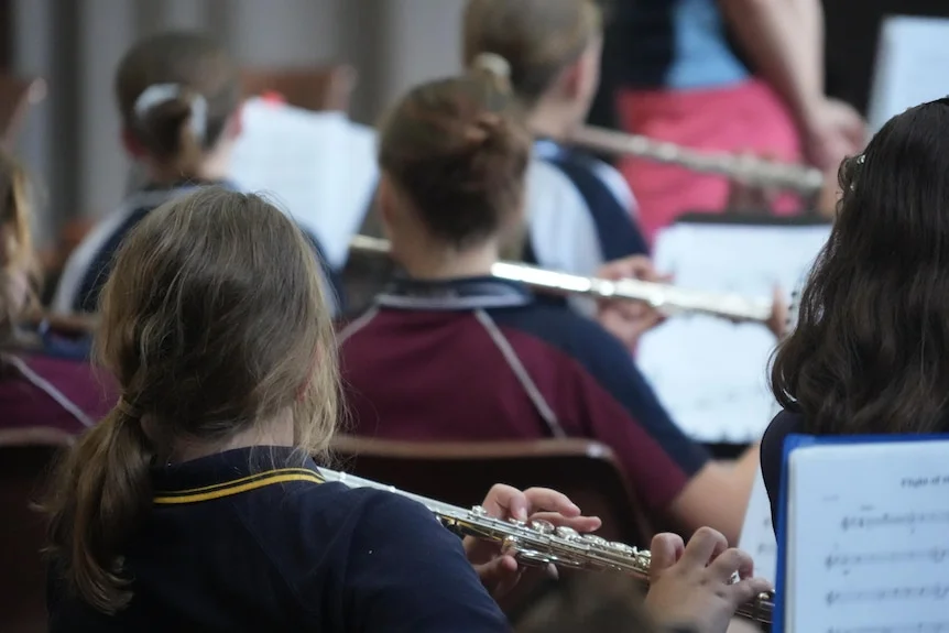 A line of young girls' heads, taken from behind, with the girls playing flutes.