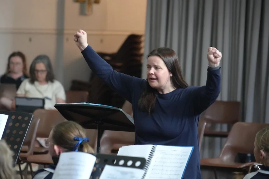 A brunette woman standing in front of a group of children playing instruments, her arms up to the sky.