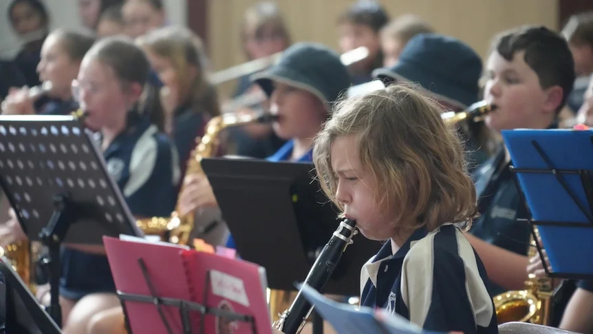 A young boy blows into a clarinet within a group of students playing instruments.