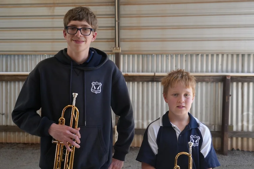 A tall teenage boy and a short young boy, both holding trumpets and smiling at the camera.