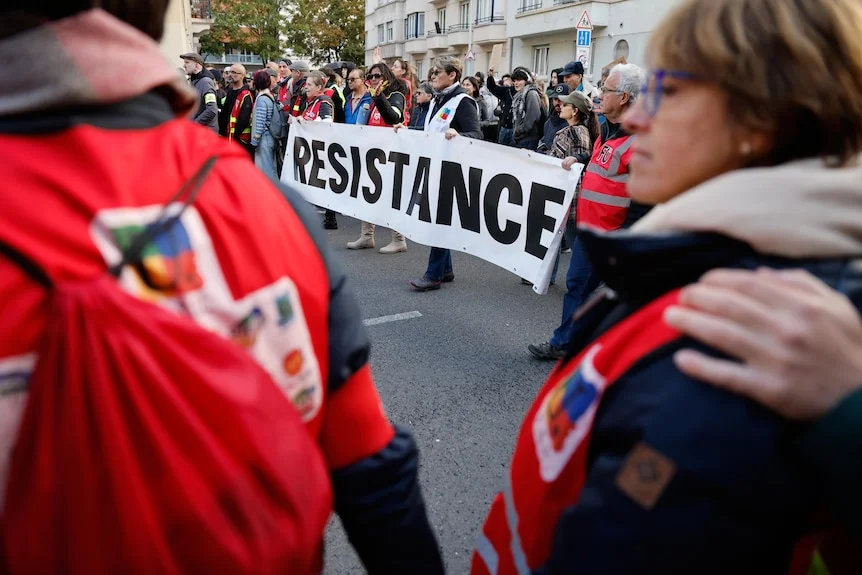 A large group of protesters walk with a white banner, with the word Resistance written in black.