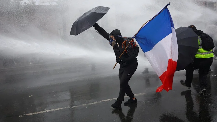 A masked protester holds a red, white and blue flag and an umbrella against a water cannon being sprayed.
