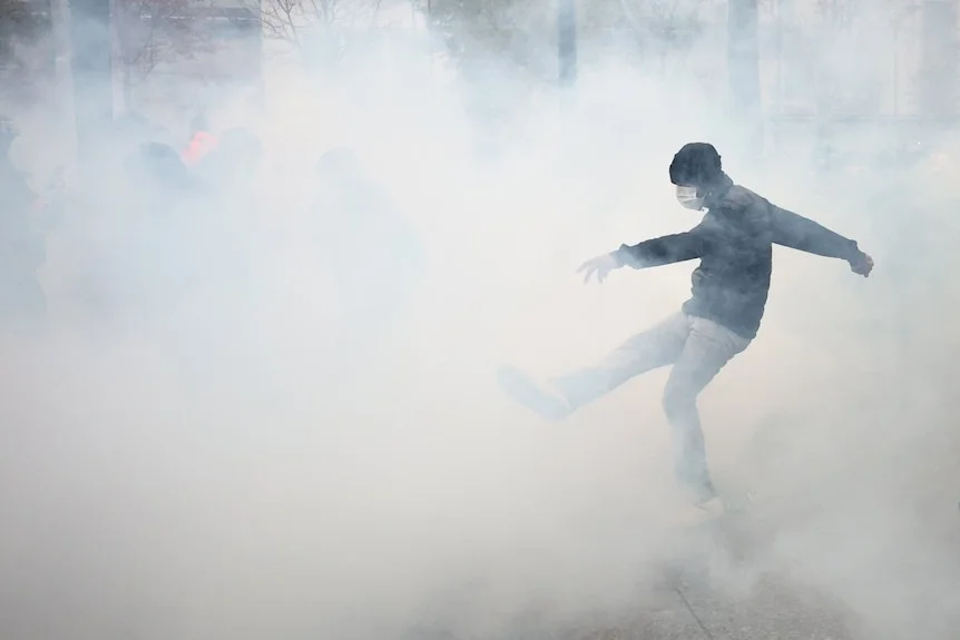 A masked protester attempts to kick a tear gas canister as smoke fills the air.