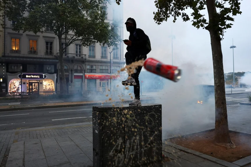 A masked protester stands on a bench and kicks a can of drink. The air is full of white smoke.