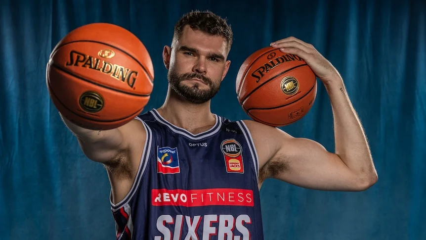Isaac Humphries in 36ers jersey holds one basketball outward and another on his other shoulder