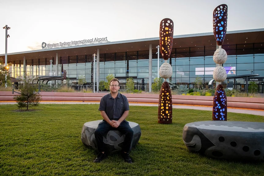 A man sitting outside an airport terminal called Western Sydney International, next to Indigenous sculture artwork, in the AM.