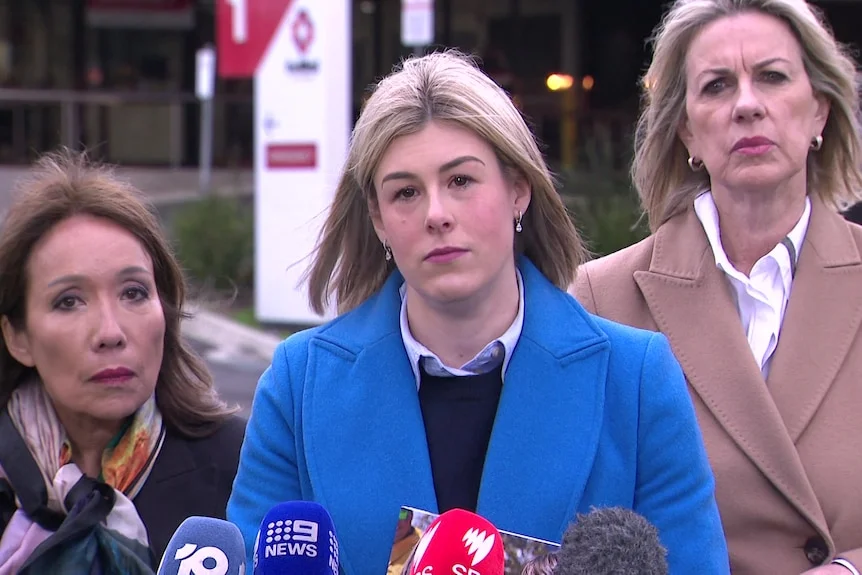 A politician, Jess Wilson, stands in front of microphones wearing a bright blue jacket.