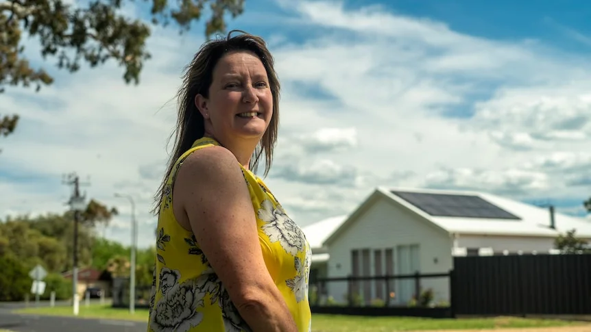 A woman in a yellow  top smiles at camera; a white house is behind her.