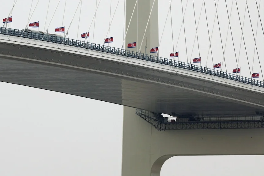 North Korean flags line the railing of a cable-stayed bridge, with buses visible on the deck.
