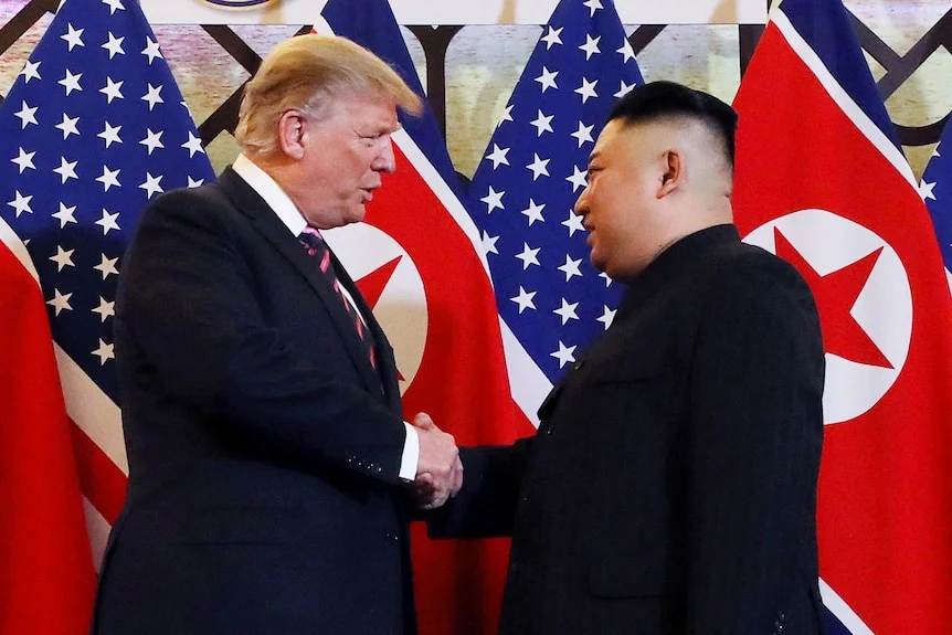 Donald Trump and Kim Jong Un in dark suits shake hands in front of alternating US and North Korean flags.