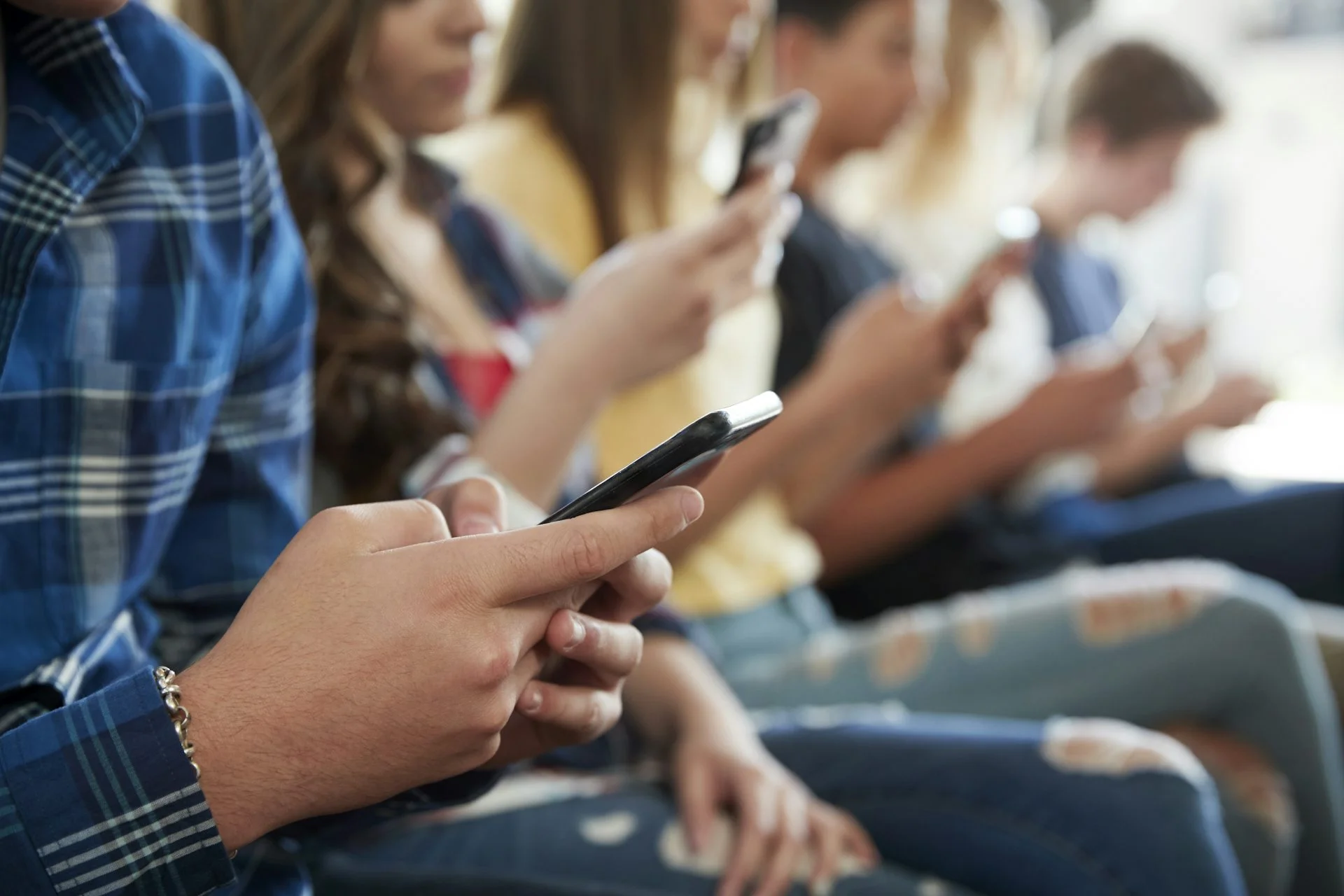 A row of teens sit next to each other while each looks at a cellphone screen