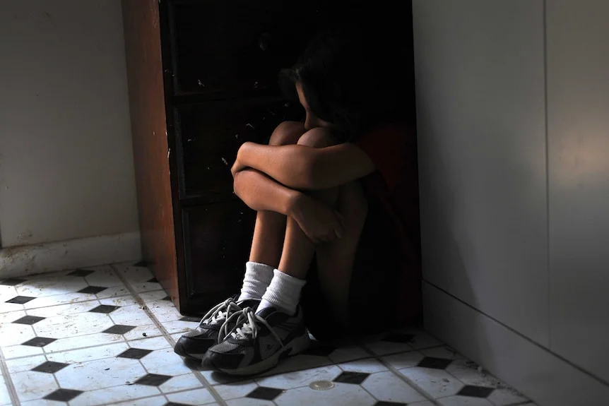 A girl sitting in a corner next to a cupboard.