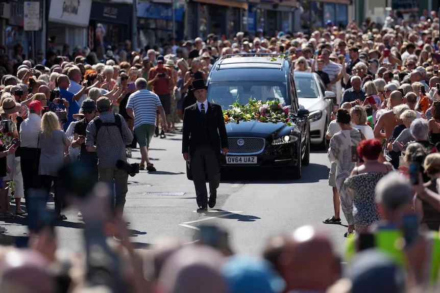 A hearse surrounded by crowds of people is guided by a man in a black suit.