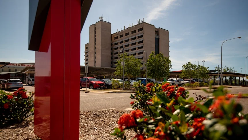 The Royal Darwin Hospital building seen from a distance, on a sunny day, with a red flower bush in the foreground.
