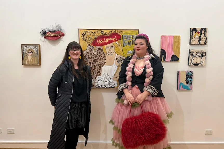 Two women stand in front of a painting in an art gallery.