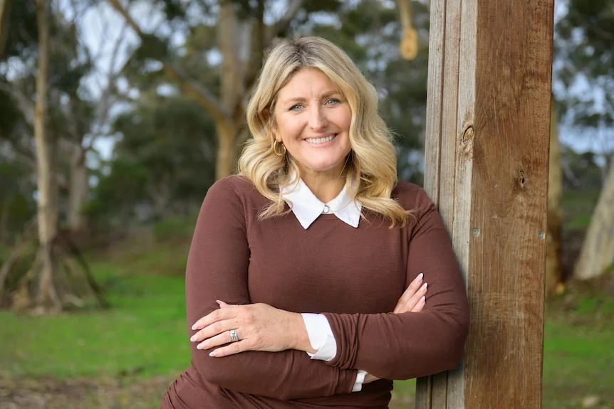 A smiling blonde woman in a brown jumper leans against a wooden post.