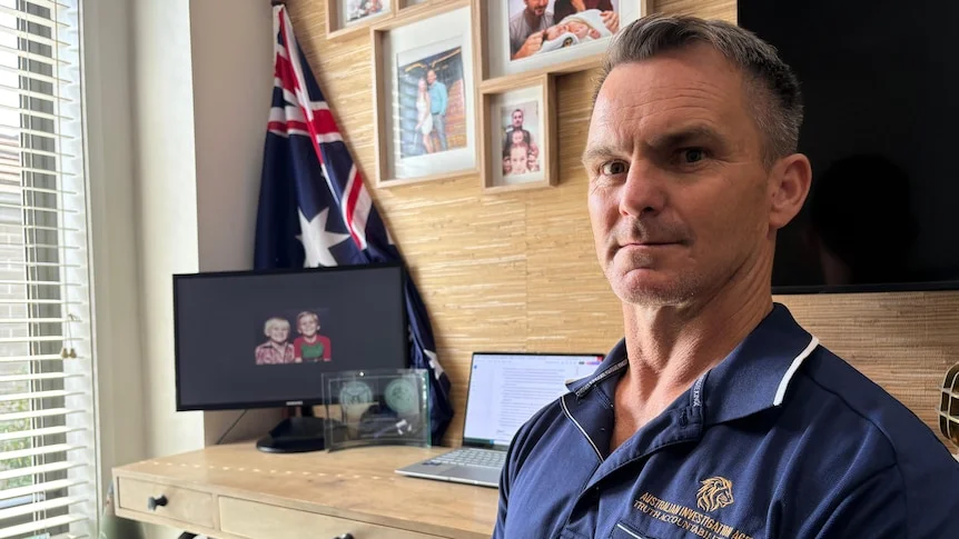 Veteran John Armfield sits by a desk with an Australian flag in the background and a photograph of him and his late brother