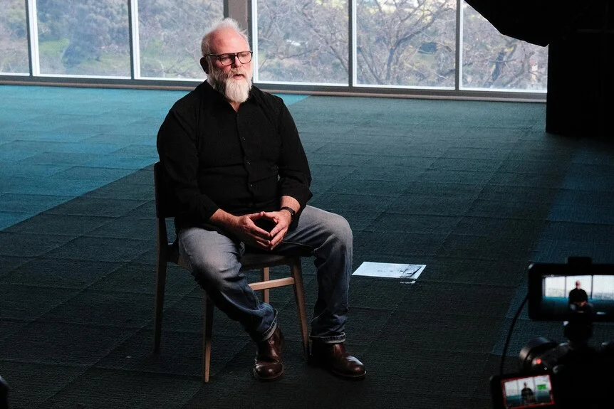 A man with white hair and a beard sits on a chair and talks to documentary makers