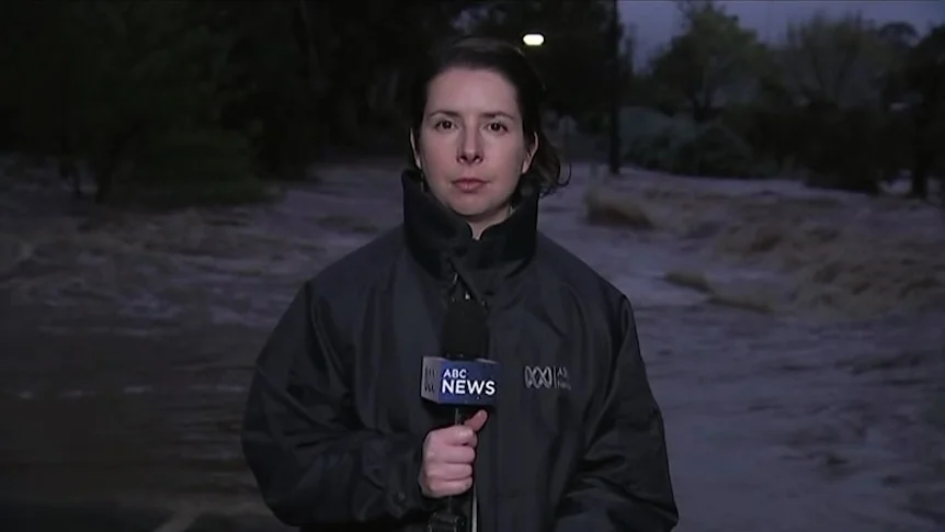 A reporter is wearing a rain jacket and holding a microphone as storm waters rage behind her.