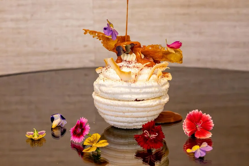 A hand pours syrup over a ball of white meringue topped with and surrounded by colourful flowers.