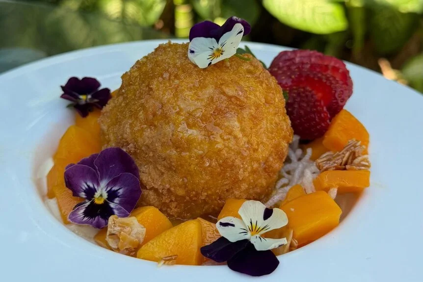 A large deep-fried scoop of ice cream sitting in a bowl, fresh fruit and small floral garnishes decorate the dish.