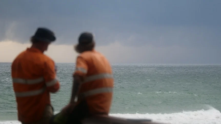 Storm clouds coming in over a beach while two men in tradie uniform look at the landscape 