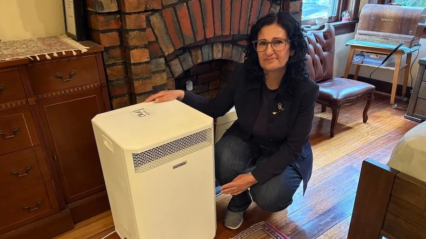 A woman kneels next to an air purifier in front of an old fireplace.