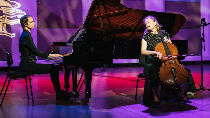 Charlotte Miles plays the cello with Laurence Matheson playing the piano to Charlotte's left on a stage lit purple