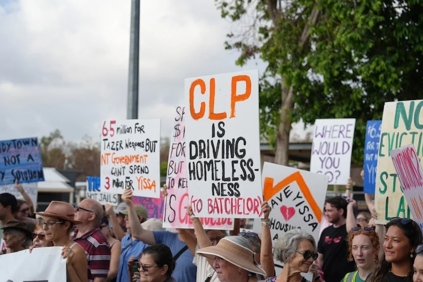A person holds a sign reading "CLP is driving homelessness (to Batchelor)" while surrounded by other protestors.