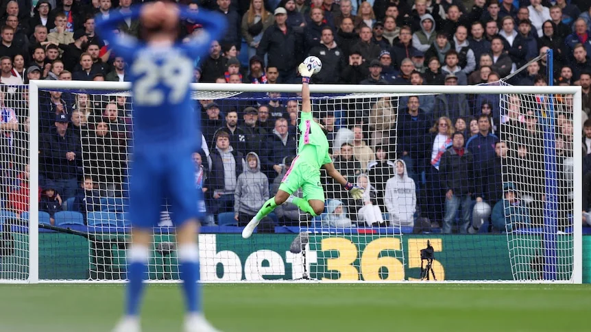 A goalkeeper dives to save shot on goal as player watches with hands on head