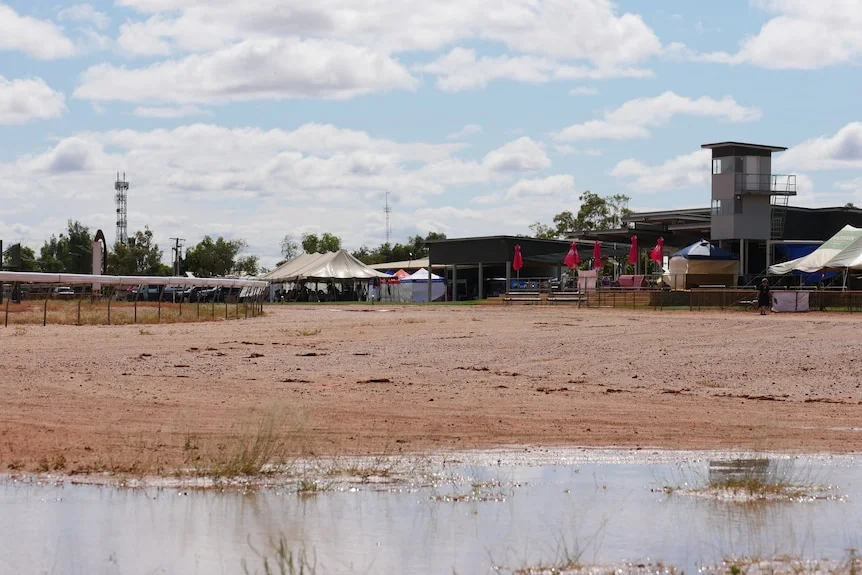 A large puddle at a red dirt racetrack under partly cloudy skies.