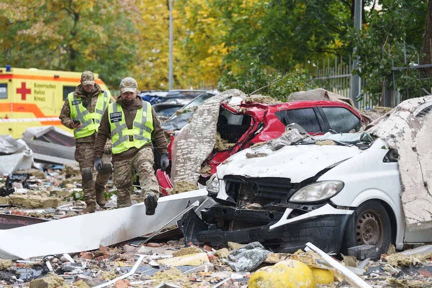 Two policemen walk in front of destroyed cars in the yard of a damaged multi-storey residential building.