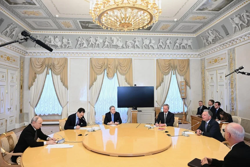 Putin, left, and Witkoff, second right, sit at a round table with four other men. Three windows are draped with gold curtains.