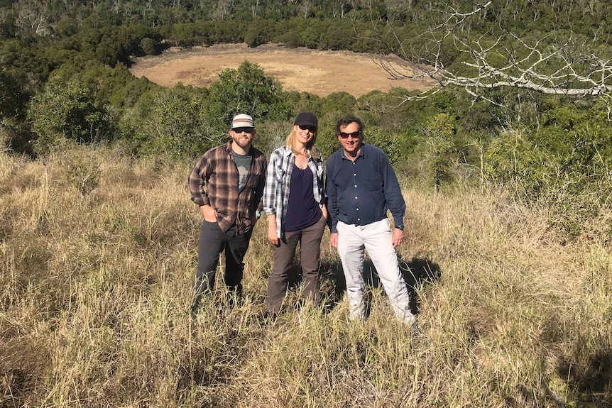 A woman with a man either side of her standing on a hill with a bush valley behind them.