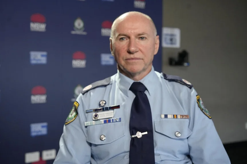 a bald man with blue eyes looks stern in police officer uniform in front of a NSW Police backdrop