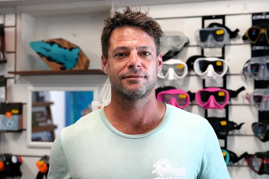 A man with short hair wearing a t-shirt looking to the camera with a slight smile. Behind him are snorkel goggles. 