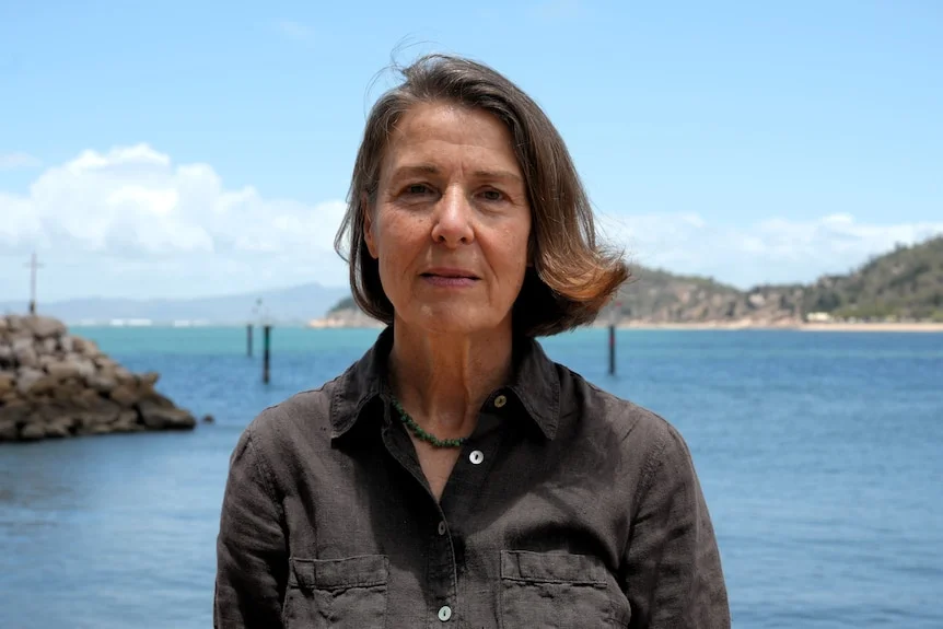A woman with short brown hair looks sternly forward. Behind her are blue waters and skies. 