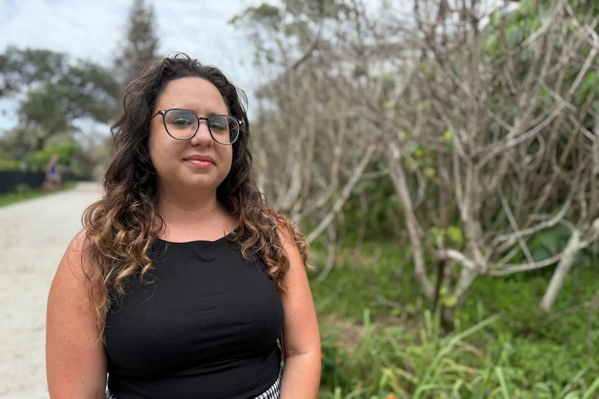 Woman with long hair, with glasses, black top, standing on path with dead trees nearby