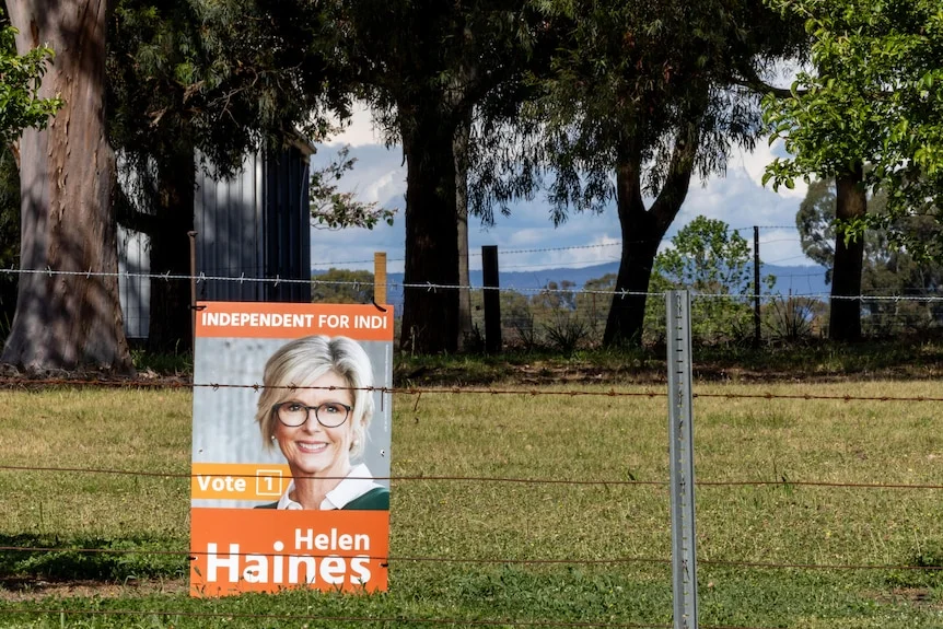A Helen Haines campaign poster affixed to a barbed-wire fence in a country area.