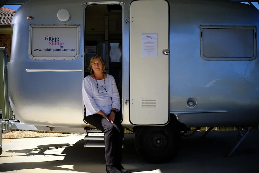 An older woman sits in the entry way of a caravan that has been fitted into a bra fitting studio.
