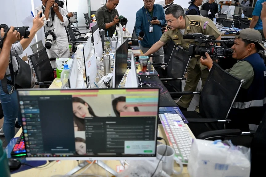 A Cambodian police officer with journalists examine a row of computers on desks. 