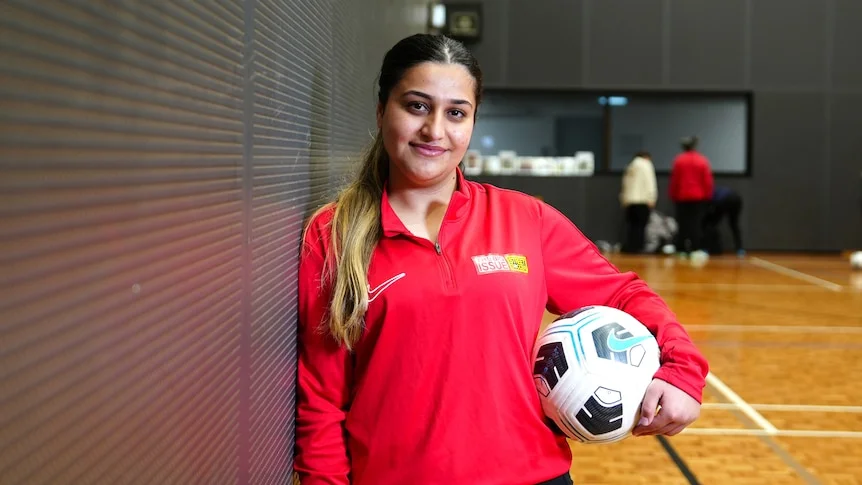 A woman wears a red jumper and holds a soccer ball out to the camera.