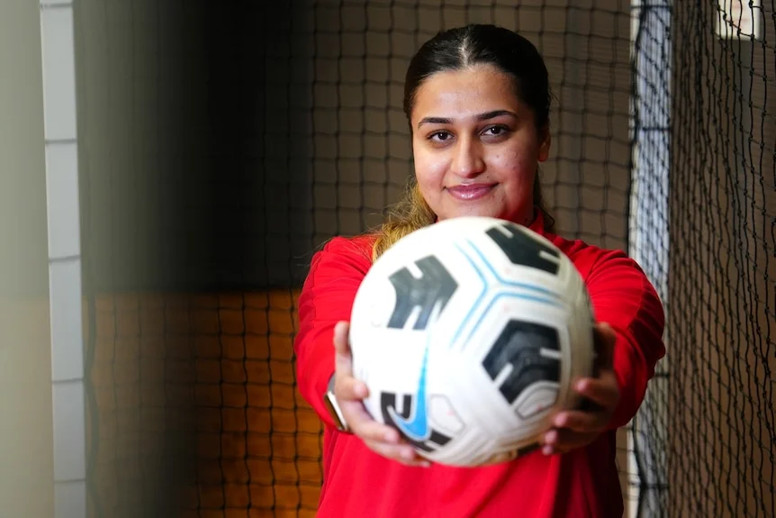A woman wears a red jumper and holds a soccer ball out to the camera.