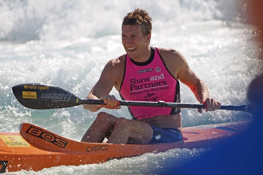 Mr Verco smiling as he uses one big oar to propel himself in a thin flat board forward in waves.