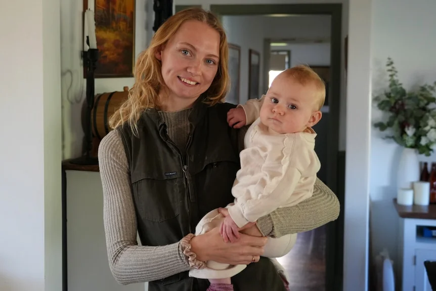 A blonde woman in a dark fleece vest and light long sleeve top looking at camera holding a baby wearing white.