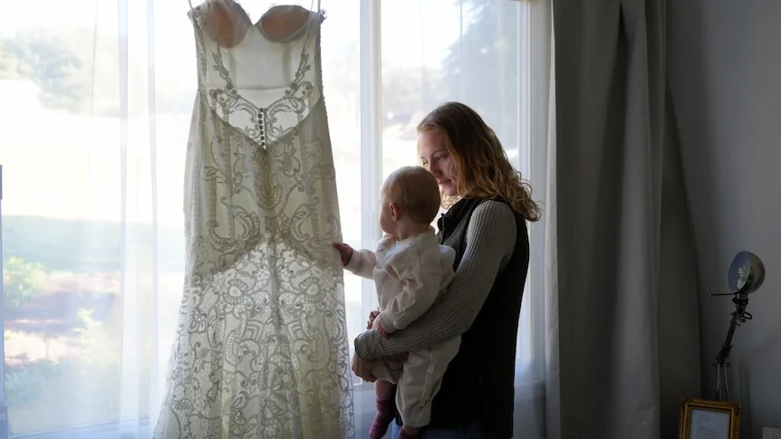 A blonde woman holding a baby by a window with a wedding dress hung in front of it with the light shining through its lace.