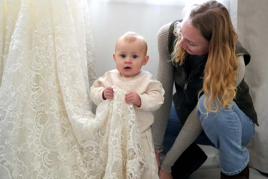 Baby wearing white standing holding the train of a lace wedding dress and looking at the camera with mum crouched beside