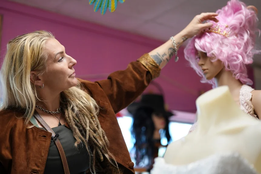 A blonde woman with dreadlocks reaching out her left hand to adjust a headband on a mannequins head.