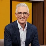 David Speers sits on a couch in front of a yellow screen, smiling at the camera, in a suit and open-necked shirt.