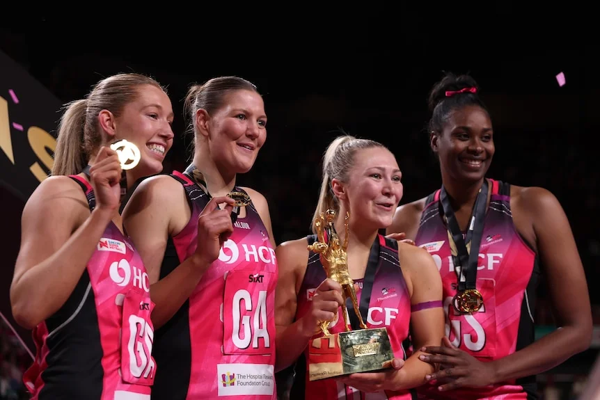 Players stand together and hold their medals and the Super Netball trophy as they smile for the camera